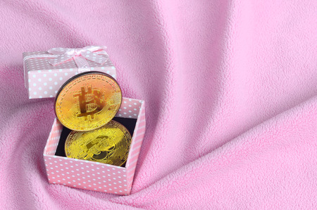 The golden bitcoin lies in a small pink gift box with a small bow on a blanket made of soft and fluffy light pink fleece fabric with a large number of relief foldsの写真素材
