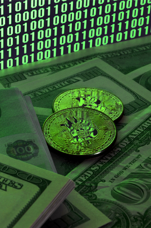 Two bitcoins lies on a pile of dollar bills on the background of a monitor depicting a binary code of bright green zeros and one units on a black background. Low key lightingの写真素材