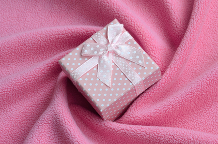 A small gift box in pink with a small bow lies on a blanket of soft and furry light pink fleece fabric with a lot of relief folds. Packing for a gift to your lovely girlfriendの写真素材
