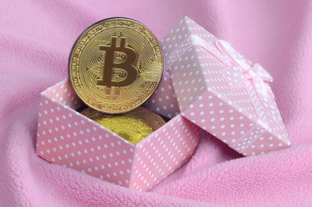 The golden bitcoin lies in a small pink gift box with a small bow on a blanket made of soft and fluffy light pink fleece fabric with a large number of relief foldsの写真素材
