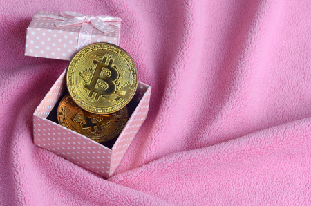 The golden bitcoin lies in a small pink gift box with a small bow on a blanket made of soft and fluffy light pink fleece fabric with a large number of relief foldsの写真素材