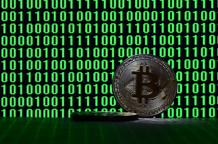 A pair of bitcoins lies on a cardboard surface on the background of a monitor depicting a binary code of bright green zeros and one units on a black background. Low key lightingの写真素材