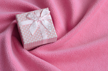 A small gift box in pink with a small bow lies on a blanket of soft and furry light pink fleece fabric with a lot of relief folds. Packing for a gift to your lovely girlfriendの写真素材