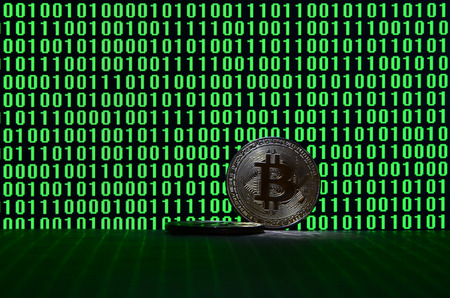 A pair of bitcoins lies on a cardboard surface on the background of a monitor depicting a binary code of bright green zeros and one units on a black background. Low key lightingの写真素材