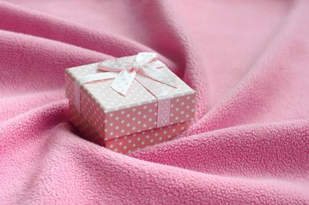 A small gift box in pink with a small bow lies on a blanket of soft and furry light pink fleece fabric with a lot of relief folds. Packing for a gift to your lovely girlfriendの写真素材