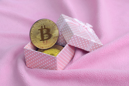 The golden bitcoin lies in a small pink gift box with a small bow on a blanket made of soft and fluffy light pink fleece fabric with a large number of relief foldsの写真素材