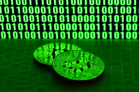 A pair of bitcoins lies on a cardboard surface on the background of a monitor depicting a binary code of bright green zeros and one units on a black background. Low key lightingの写真素材