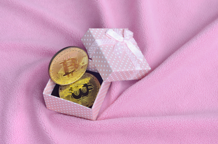 The golden bitcoin lies in a small pink gift box with a small bow on a blanket made of soft and fluffy light pink fleece fabric with a large number of relief foldsの写真素材