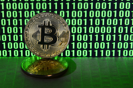 A pair of bitcoins lies on a cardboard surface on the background of a monitor depicting a binary code of bright green zeros and one units on a black background. Low key lightingの写真素材