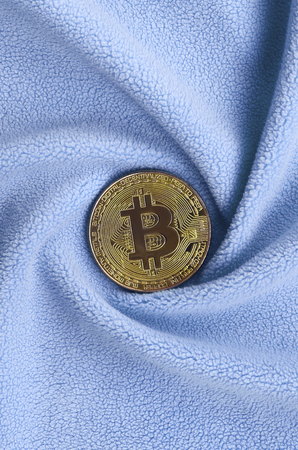 The golden bitcoin lies on a blanket made of soft and fluffy light blue fleece fabric with a large...