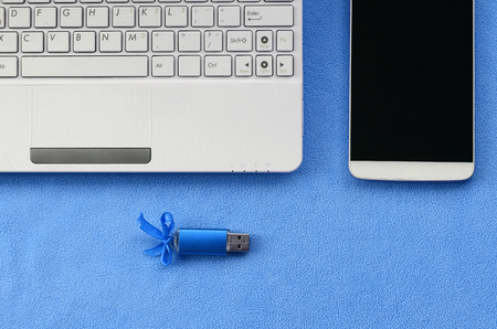 Brilliant blue usb flash memory card with a blue bow lies on a blanket of soft and furry light blue fleece fabric beside to a white laptop and smartphone. Classic female gift design for a memory cardの写真素材