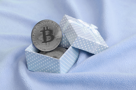 The silver bitcoin lies in a small blue gift box with a small bow on a blanket made of soft and fluffy light blue fleece fabric with a large number of relief foldsの写真素材