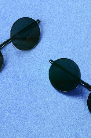 Two stylish black sunglasses with round glasses lies on a blanket made of soft and fluffy light blue fleece fabric. Fashionable background picture in female colorsの写真素材