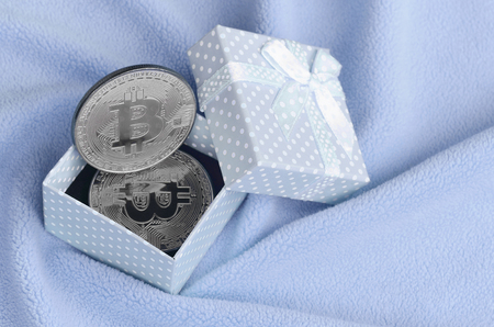 The silver bitcoin lies in a small blue gift box with a small bow on a blanket made of soft and fluffy light blue fleece fabric with a large number of relief foldsの写真素材