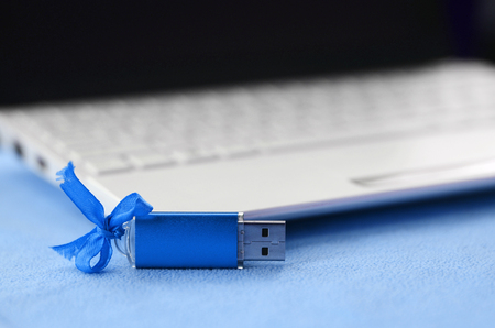 Brilliant blue usb flash memory card with a blue bow lies on a blanket of soft and furry light blue fleece fabric beside to a white laptop. Classic female gift design for a memory cardの写真素材
