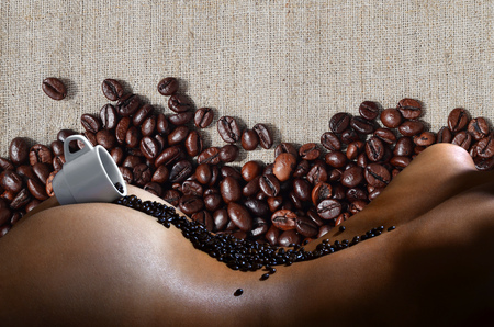 A photograph of a white cup of hot coffee with lots of coffee beans, which lies on a beautiful and sexy naked female body with a many beans in background pattern. Erotic photo about the coffee drinksの写真素材