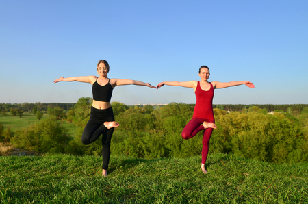 Two young fair-haired girls in sports suits practice yoga on a picturesque green hill in the open air in the evening. The concept of sport  exercising and healthy lifestylesの写真素材