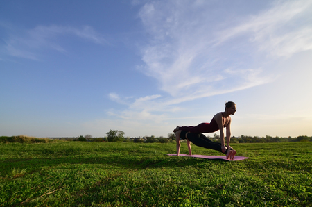 Two young fair-haired girls in sports suits practice yoga on a picturesque green hill in the open air in the evening. The concept of sport  exercising outdoorsの写真素材