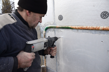 An elderly workman drills a hole in a styrofoam wall for the subsequent installation of a plastic reinforcing dowel. Creating holes in the wall with a drill. Warming of the building facadeの写真素材