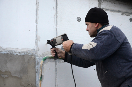 An elderly workman drills a hole in a styrofoam wall for the subsequent installation of a plastic reinforcing dowel. Creating holes in the wall with a drill. Warming of the building facadeの写真素材
