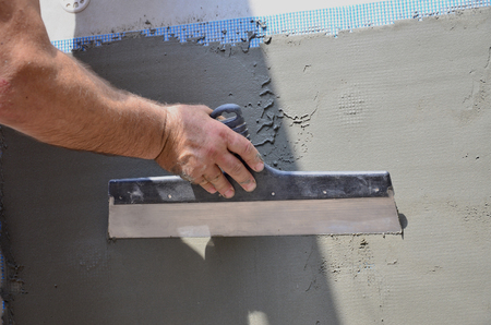 Hands of an old manual worker with wall plastering tools renovating house. Plasterer renovating outdoor walls and corners with spatula and plaster. Wall insulation. Construction finishing worksの写真素材