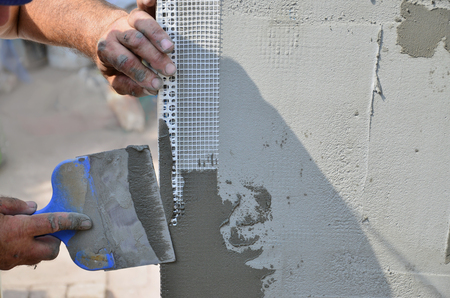 Hands of an old manual worker with wall plastering tools renovating house. Plasterer renovating outdoor walls and corners with spatula and plaster. Wall insulation. Construction finishing worksの写真素材