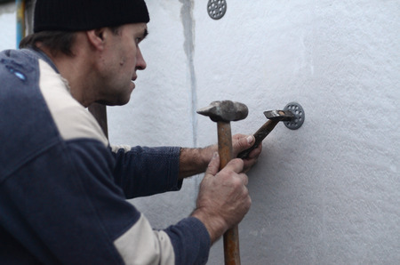 An elderly workman clogs a dowel into a plastic umbrella mount in a styrofoam wall. The process of fixing expanded polystyrene plates. Warming of the facade of the buildingの写真素材