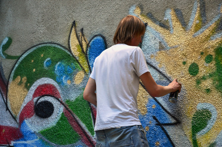 Photo in the process of drawing a graffiti pattern on an old concrete wall. Young long-haired blond guy draws an abstract drawing of different colors. Street art and vandalism conceptの写真素材