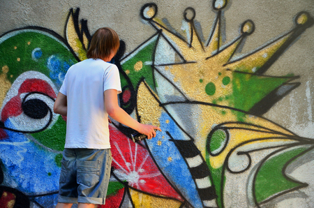 Photo of a young guy in denim shorts and a white shirt. The guy draws on the graffiti wall a drawing with aerosol paints of various colors. The concept of hooliganism and damage to propertyの写真素材
