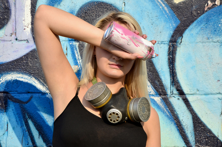 Young and beautiful sexy girl graffiti artist with gas mask on her neck hiding his eyes with a spray can standing on a wall background with a graffiti pattern in blue and purple tonesの写真素材