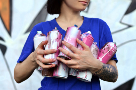 Portrait of an emotional young girl with black hair and piercings. Photo of a girl with aerosol paint cans in hands on a graffiti wall background. The concept of street art and use of aerosol paintsの写真素材