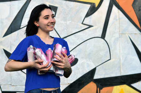 Portrait of an emotional young girl with black hair and piercings. Photo of a girl with aerosol paint cans in hands on a graffiti wall background. The concept of street art and use of aerosol paintsの写真素材