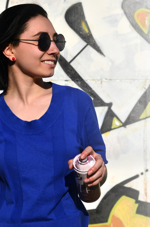 Portrait of an emotional young girl with black hair and piercings. Photo of a girl with aerosol paint cans in hands on a graffiti wall background. The concept of street art and use of aerosol paintsの写真素材