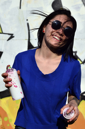 Portrait of an emotional young girl with black hair and piercings. Photo of a girl with aerosol paint cans in hands on a graffiti wall background. The concept of street art and use of aerosol paintsの写真素材