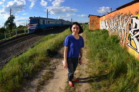 Portrait of an emotional young girl with black hair and piercings. A wide-angle photo of a girl with aerosol paint cans in the hands on a graffiti wall background. A modern portrait of a fisheye lensの写真素材