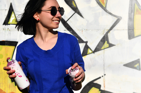 Portrait of an emotional young girl with black hair and piercings. Photo of a girl with aerosol paint cans in hands on a graffiti wall background. The concept of street art and use of aerosol paintsの写真素材
