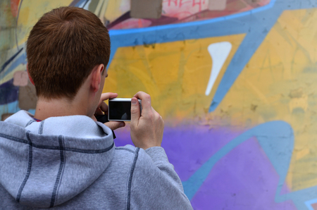 A young graffiti artist photographs his completed picture on the wall. The guy uses modern technology to capture a colorful abstract graffiti drawing. Focus on the photographing deviceの写真素材