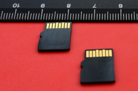 Two small micro SD memory cards lie on a red background next to a black ruler. A small and compact data and information storeの写真素材