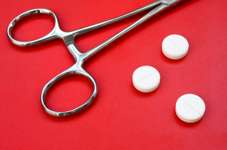 Several white tablets and a medical clamp lie on a bright red background surface. Background image on medical and pharmaceutical topicsの写真素材