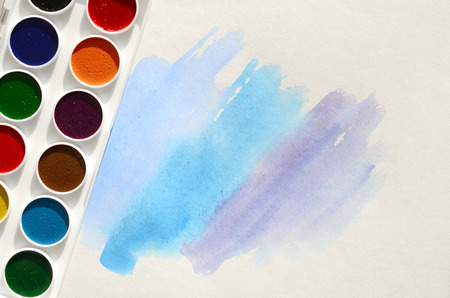 A new set of watercolors lies on a sheet of paper, which shows an abstract watercolor drawing in the form of blue strokes. The concept of amateur painting among teenagersの写真素材
