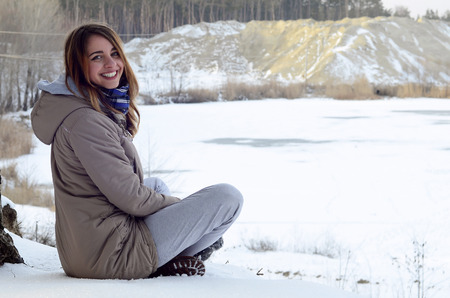 A young Caucasian girl in a brown coat is sitting near a cliff in the background of a horizon line between the sky and a frozen lake in winter timeの写真素材