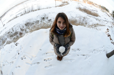 A young and joyful Caucasian girl in a brown coat holds a snowball in front of a horizon line...