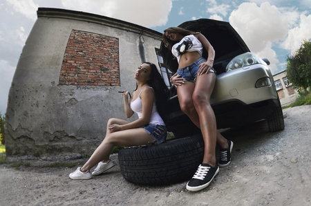 Two young and sexy caucasian girls (blonde and brunette) with wheel wrenches around a big silvery off-road car with an open hood. The concept of repair and maintenance of a carの写真素材