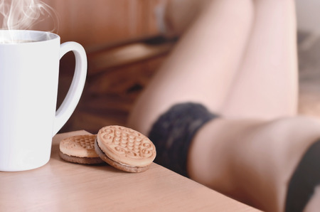 A cup of hot coffee and round cookies with a silhouette of a sexy girl in black underwear in the background on a bed. Focus foreground priority. Small white coffee cup with steam and brown biscuits の写真素材