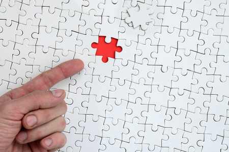 The texture of a white jigsaw puzzle in the assembled state with one missing element, forming a red space, pointed to by the finger of the male handの写真素材
