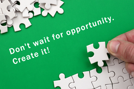 Don't wait for opportunity. Create it. The hand folds a white jigsaw puzzle and a pile of uncombed puzzle pieces lies against the background of the green surfaceの写真素材