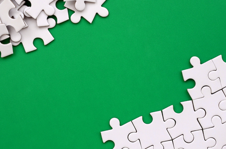 Fragment of a folded white jigsaw puzzle and a pile of uncombed puzzle elements against the background of a green surface. Texture photo with space for textの写真素材