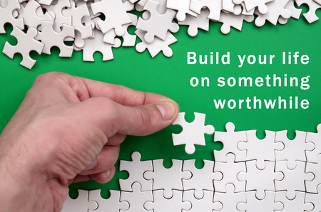 Build your life on something worthwhile. The hand folds a white jigsaw puzzle and a pile of uncombed puzzle pieces lies against the background of the green surfaceの写真素材