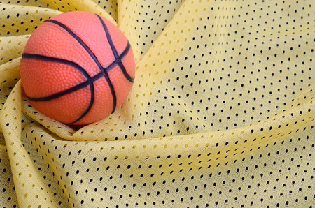 Small orange rubber basketball lies on a yellow sport jersey clothing fabric texture and background with many foldsの写真素材
