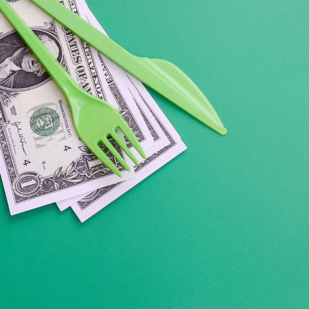 Disposable plastic cutlery green. Plastic fork and knife lie on a small amount of US dollarsの写真素材